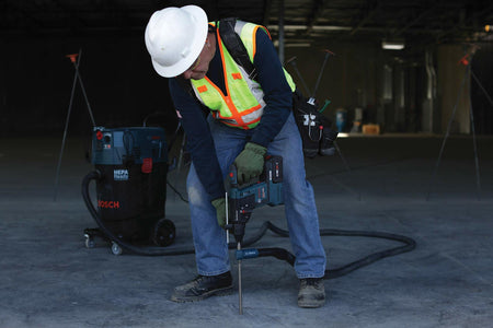 Worker holding Bosch rotary hammer drill connected to HEPA-ready vacuum system. Wearing safety gear and tool belt in large indoor construction site with lighting and tripods in background.