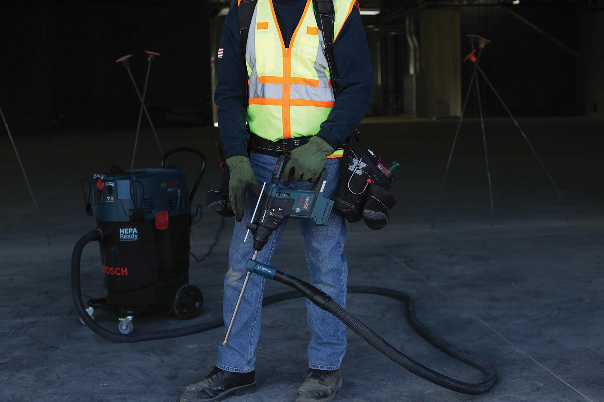 Worker holding Bosch rotary hammer drill connected to HEPA-ready vacuum system. Wearing safety gear and tool belt in large indoor construction site with lighting and tripods in background.