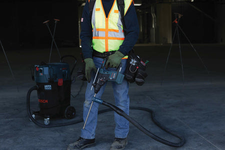 Worker holding Bosch rotary hammer drill connected to HEPA-ready vacuum system. Wearing safety gear and tool belt in large indoor construction site with lighting and tripods in background.
