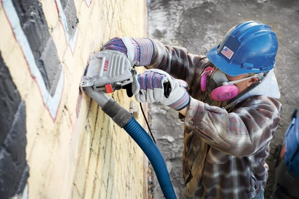 A professional mason wearing a hard hat, respirator, and gloves using a BOSCH angle grinder equipped with a DD4510S diamond sandwich blade and dust extraction vacuum to remove mortar from a brick wall.