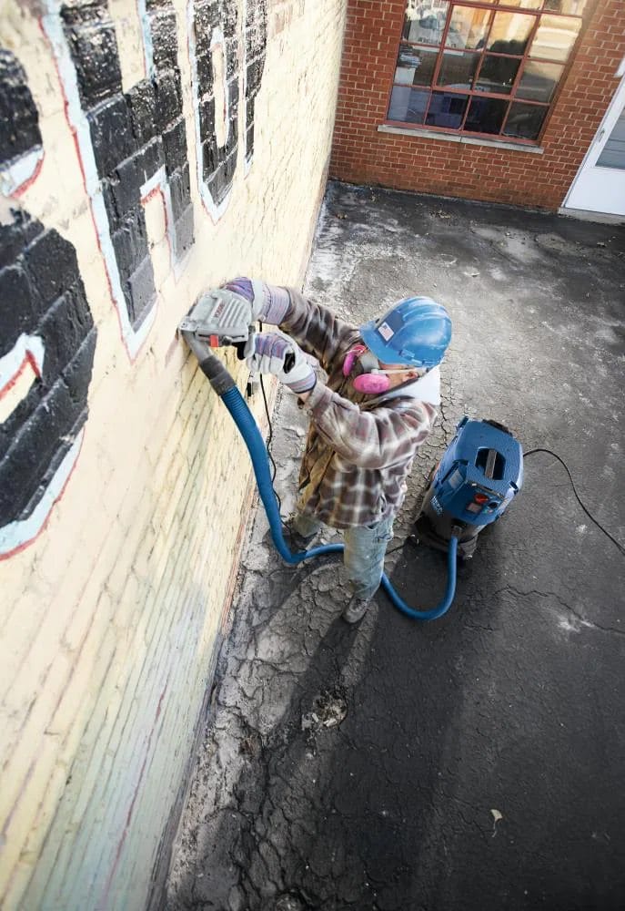 Top-down view of a technician performing masonry restoration on a brick exterior using a BOSCH 4-1/2 inch sandwich tuckpointing blade connected to a HEPA-ready dust extractor for clean grout removal.