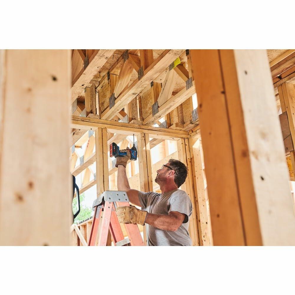 A construction worker on a ladder, using the Bosch GDR18V-1860C impact driver overhead to fasten lumber in a newly framed wooden structure.