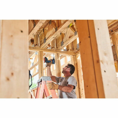 A construction worker on a ladder, using the Bosch GDR18V-1860C impact driver overhead to fasten lumber in a newly framed wooden structure.