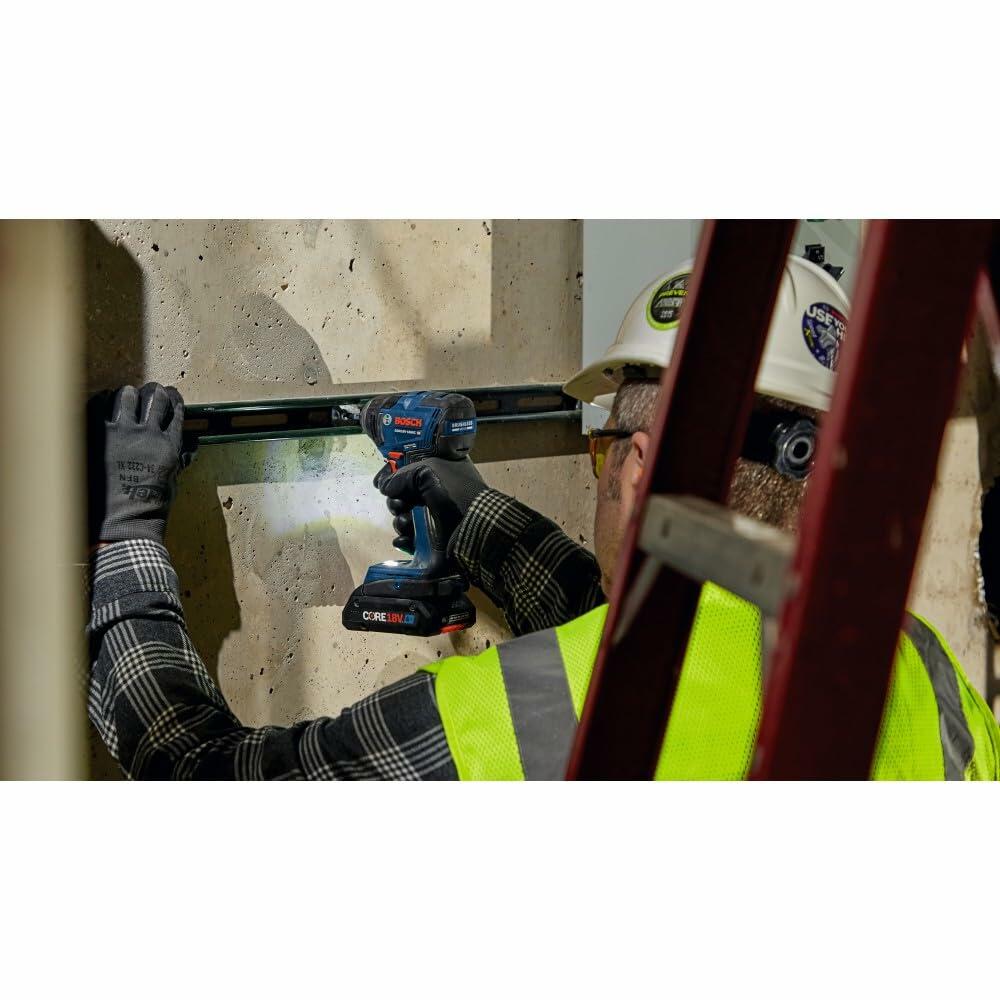 A construction worker wearing a hard hat and safety vest, using the Bosch GDR18V-1860C impact driver to fasten material to a concrete wall.