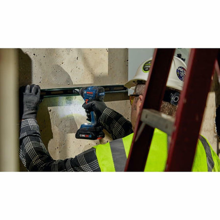 A construction worker wearing a hard hat and safety vest, using the Bosch GDR18V-1860C impact driver to fasten material to a concrete wall.