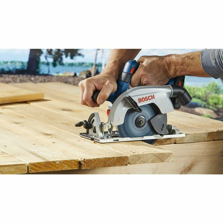A person holds the Bosch cordless circular saw with both hands, making a cross-cut on a wide wooden board outdoors.