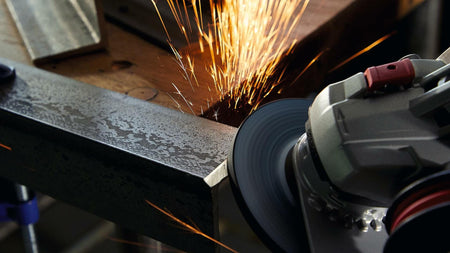 Close-up of angle grinder smoothing edge of clamped metal piece. Bright sparks fly from grinding point. Workshop setting emphasizes active metal shaping process.