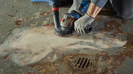 Worker using Bosch grinder with abrasive disc to remove paint from concrete floor. Vacuum hose attached for dust control. Gloves worn for safety near embedded metal floor drain