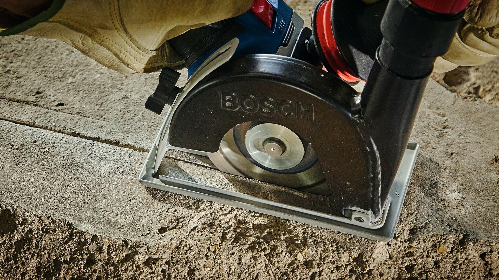 Person using Bosch angle grinder with diamond blade to cut into concrete. Tool is equipped with dust extraction attachment. Operator wears gloves for safety during outdoor construction.