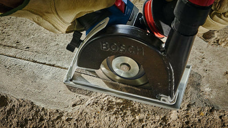 Person using Bosch angle grinder with diamond blade to cut into concrete. Tool is equipped with dust extraction attachment. Operator wears gloves for safety during outdoor construction.