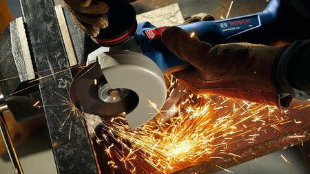 Bosch GWS13-50 angle grinder cutting metal in vise, producing bright sparks. Operator wears gloves. Scene highlights intensity and precision of metalworking task.