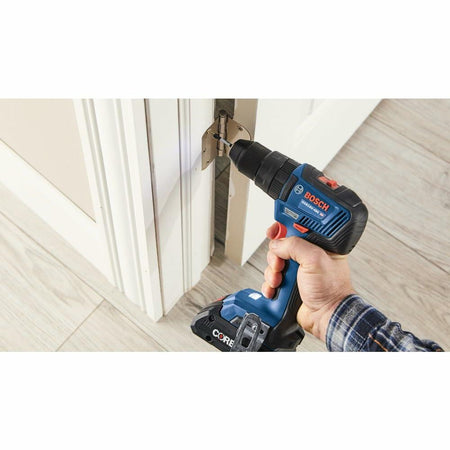 Person using a Bosch cordless power drill to install or remove a hinge on a white door frame. The drill features a visible CORE18V battery pack and is part of the Bosch GXL18V-233B25 2-tool combo kit. The user is wearing a plaid shirt and working on light wood flooring. This image demonstrates the hammer drill’s practical use in home improvement and construction tasks.