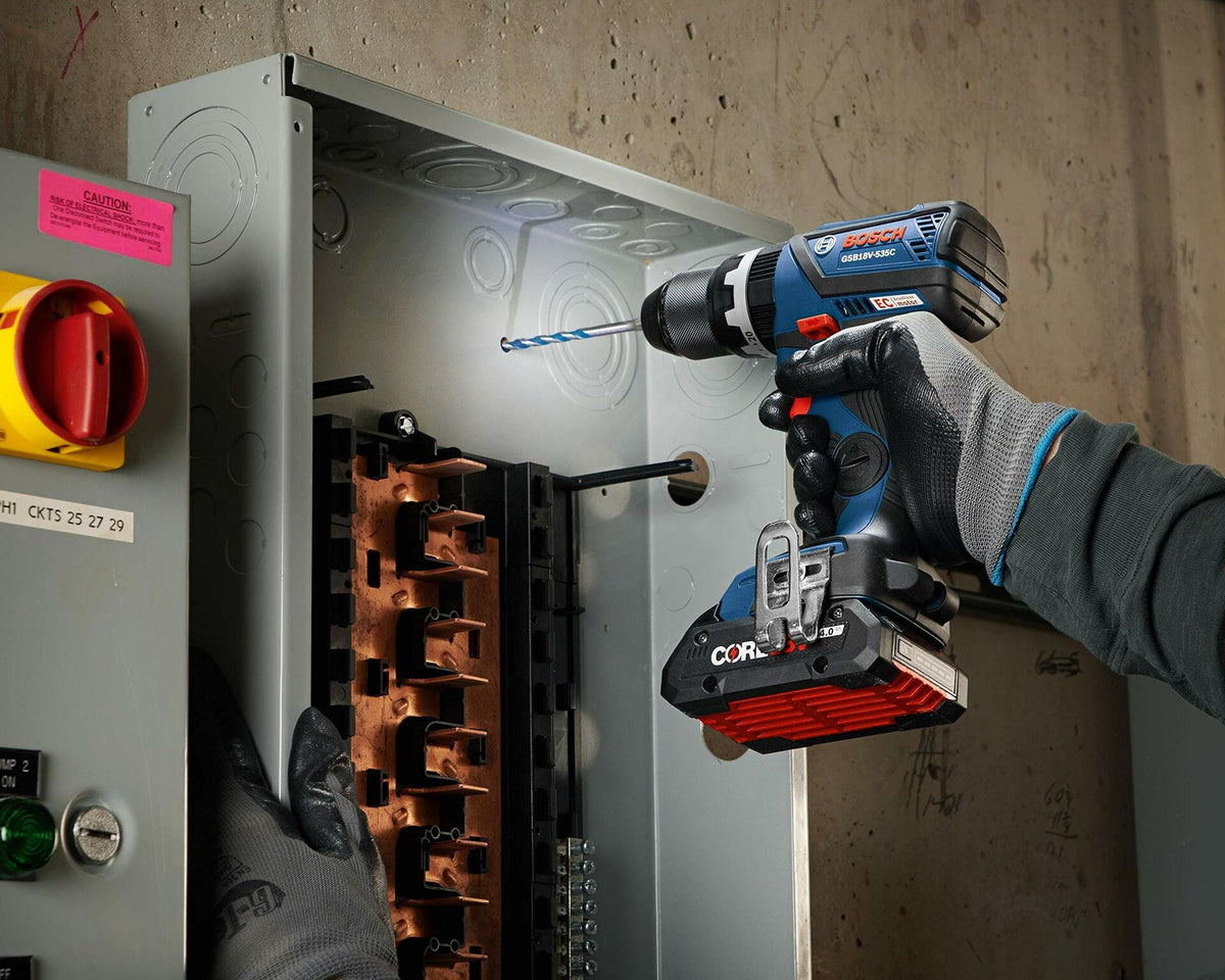 Person wearing gloves using a Bosch CORE18V cordless drill to drill into the side of an open electrical panel. Copper bus bars and internal components are visible. Scene demonstrates safe electrical work using professional-grade tools.