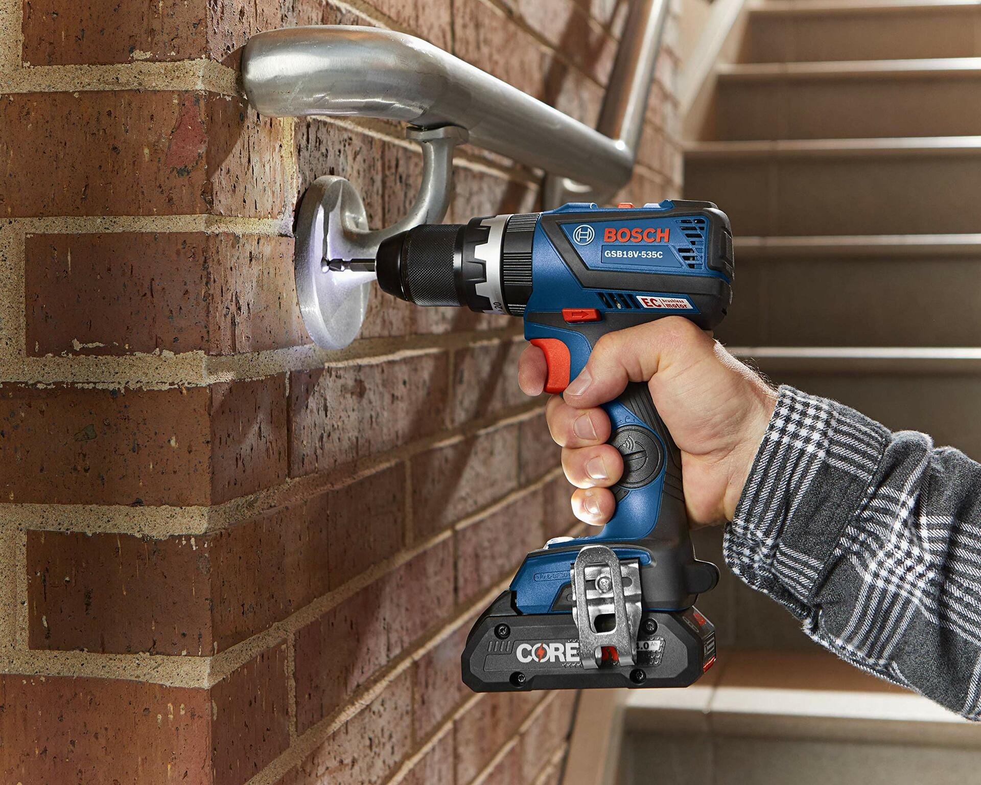 Person using a Bosch GSR18V-535C cordless drill to install a metal handrail bracket into a brick wall. Drill is equipped with a CORE18V battery. Scene highlights tool’s capability for drilling into metal and masonry surfaces during stairway construction.