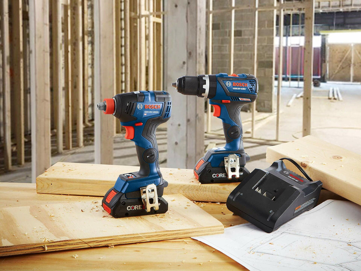 Bosch cordless impact driver and drill/driver with CORE18V batteries displayed on a wooden surface at a construction site. A Bosch battery charger and blueprint are also visible. Background includes exposed framing and concrete walls, indicating active building work.