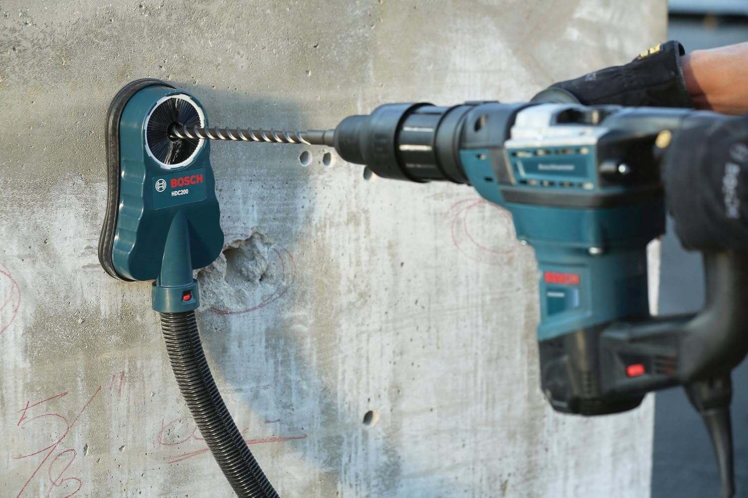 Person drilling into a concrete wall using Bosch power drill and HDC200 dust collection attachment connected to a vacuum hose; red wall markings visible near drill site.