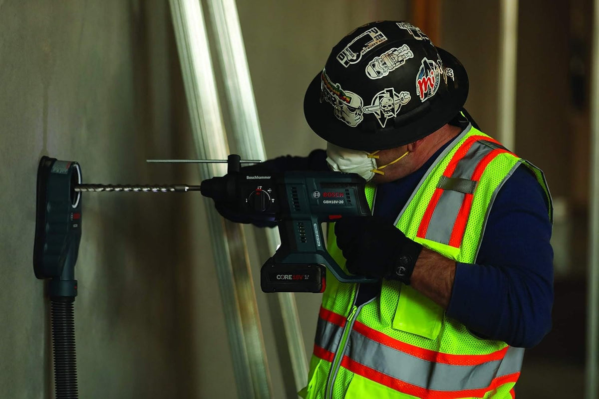 Construction worker wearing safety gear using Bosch GBH18V-20 rotary hammer drill with dust extraction system to drill into concrete wall in indoor jobsite setting.
