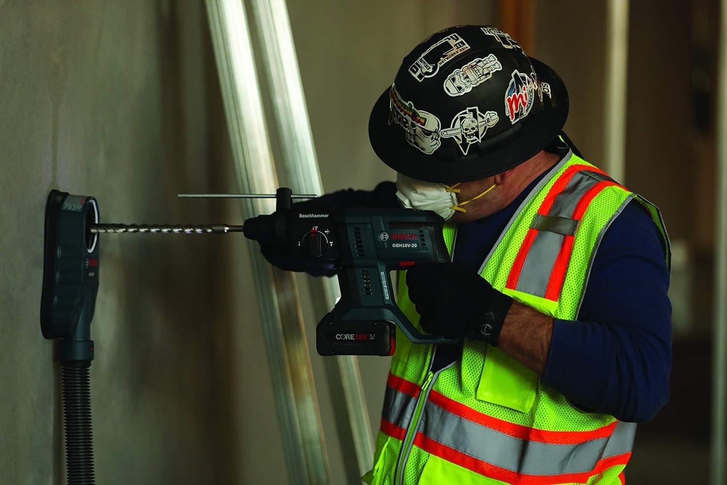 Construction worker wearing safety gear using Bosch GBH18V-20 rotary hammer drill with dust extraction system to drill into concrete wall in indoor jobsite setting.