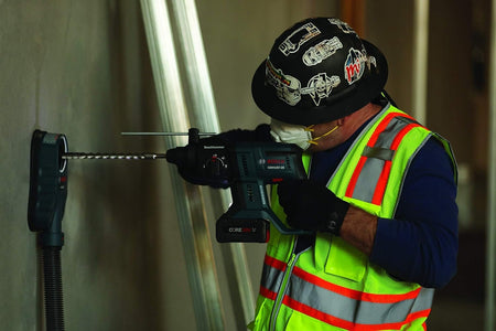 Construction worker wearing safety gear using Bosch GBH18V-20 rotary hammer drill with dust extraction system to drill into concrete wall in indoor jobsite setting.