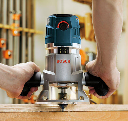 Bosch MRF23EVS router in use on a wood panel. The user grips both black handles while the cutting bit engages the surface. Workshop tools and clamps are visible in the background, indicating a professional setting.