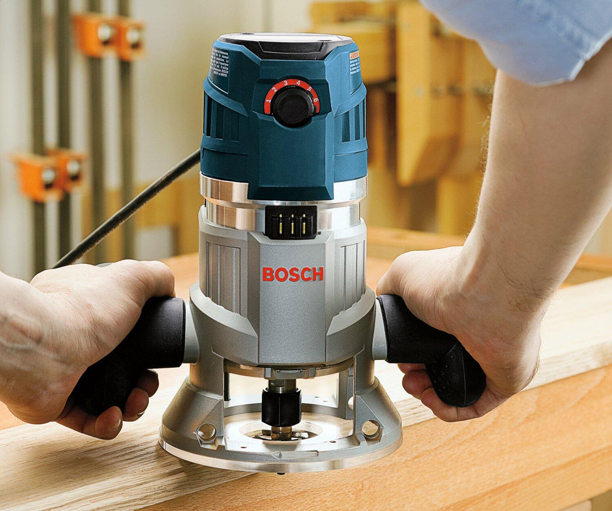 Person using Bosch MRF23EVS router on a wooden surface. The blue and silver tool is guided along the edge with both hands on black handles. The red dial and cutting bit are visible, emphasizing control and precision.