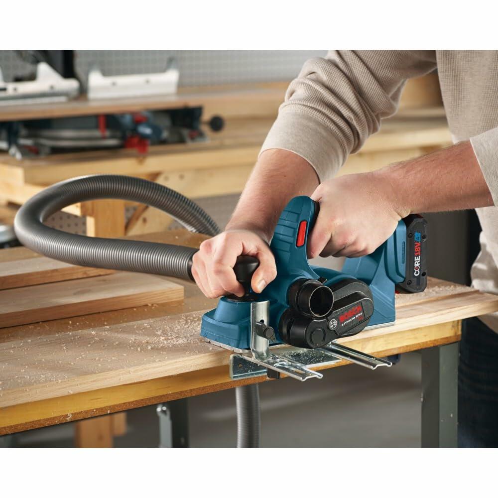 A woodworker using the Bosch cordless planer on a board in a workshop, with a dust collection hose attached to the chip-ejection port.