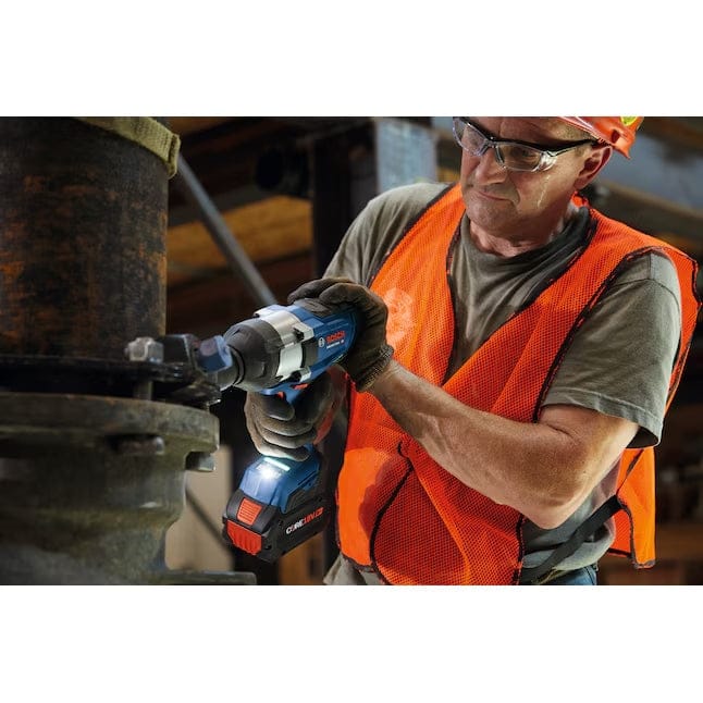 A worker in an orange vest, hard hat, and safety glasses uses the Bosch GDS18V-770CN impact wrench, with its LED light illuminating a large bolt on a piece of industrial machinery.