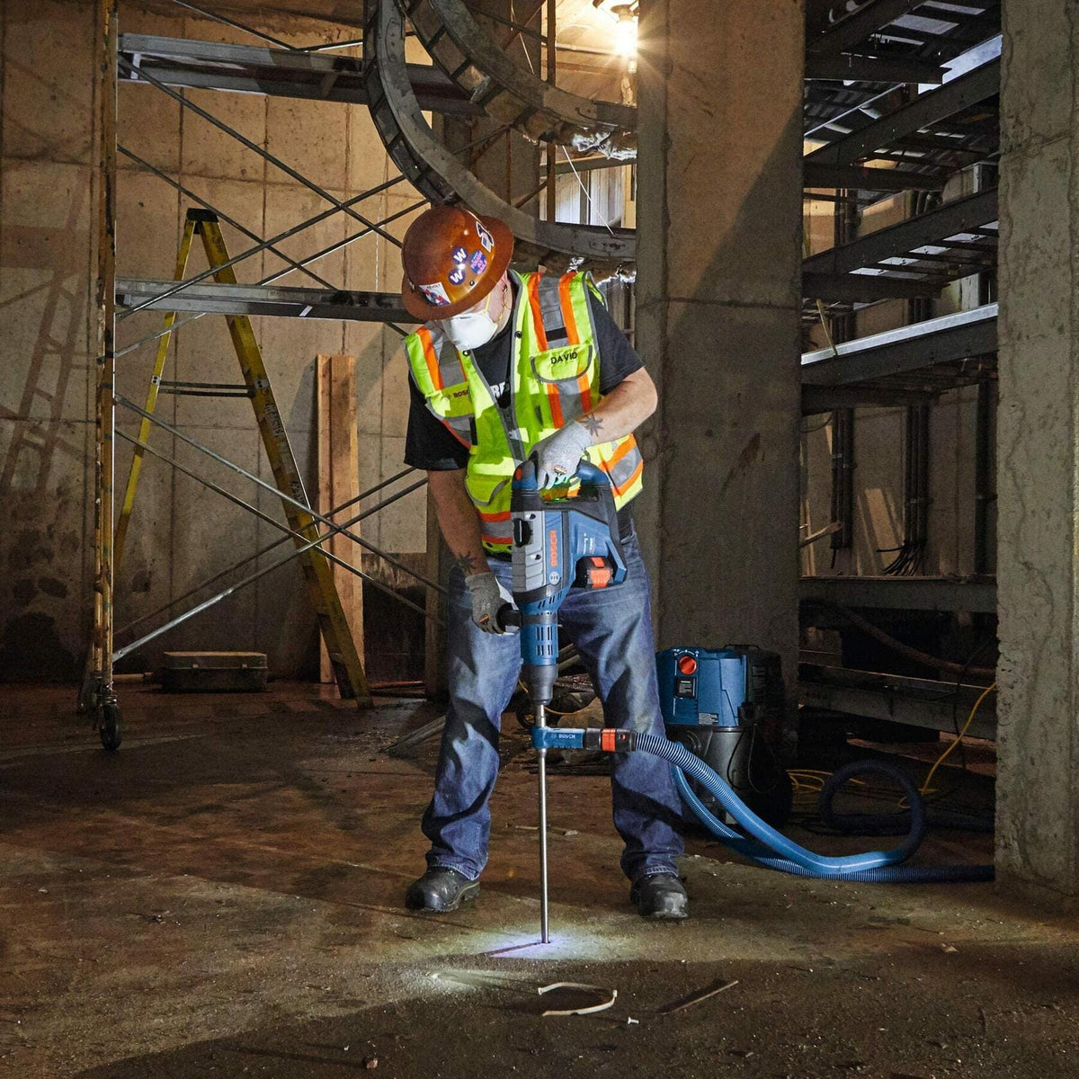 A construction worker in full safety gear uses the Bosch rotary hammer with a long drill bit to drill a hole downward into a concrete floor on an indoor jobsite.
