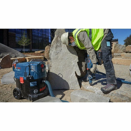 A worker in full protective gear uses the Bosch rotary hammer to drill into a large boulder while it is connected to a Bosch HEPA Ready dust extractor.