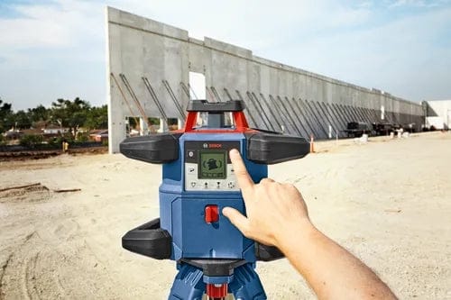 A worker manually adjusting the control panel of the Bosch REVOLVE4000 rotary laser mounted on a tripod at a large, sunny construction site with concrete walls in the background.