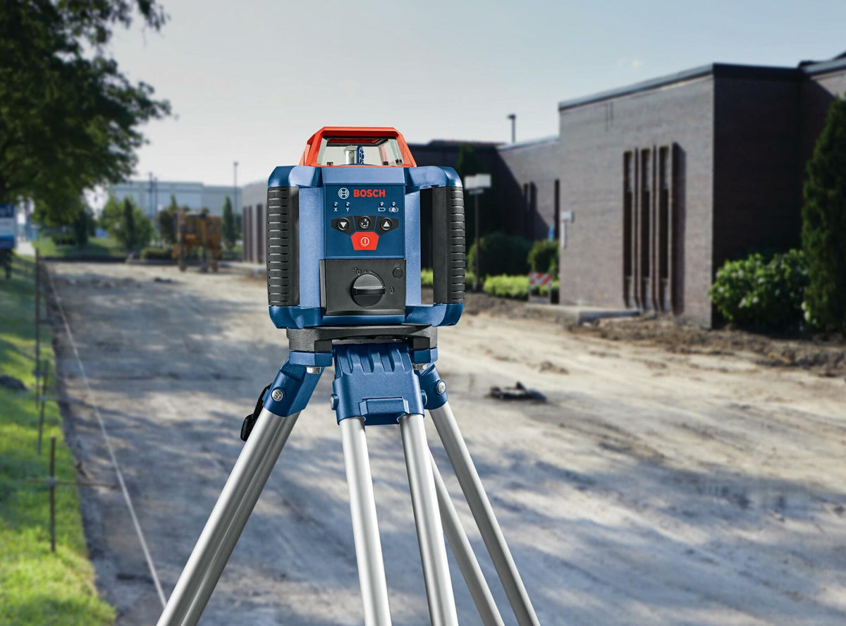 Bosch rotary laser level mounted on a tripod at a construction site. Background shows a partially paved path, construction equipment, and a brick building, emphasizing the tool’s role in precise alignment.