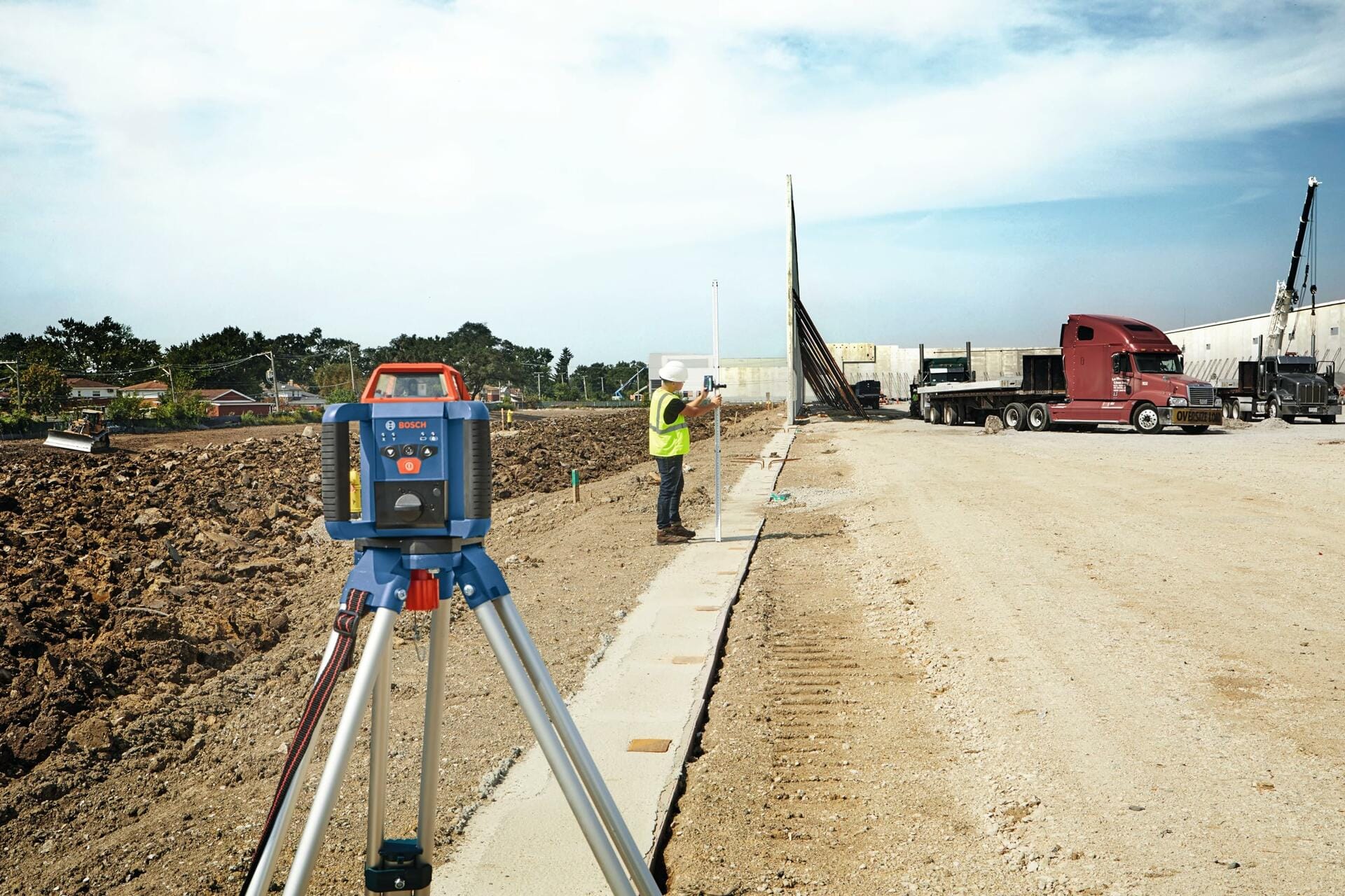 Bosch rotary laser level on a tripod in the foreground of a construction site. A worker in safety gear uses a measuring rod in the background. The site includes dirt, gravel, vehicles, and partially constructed curbs.