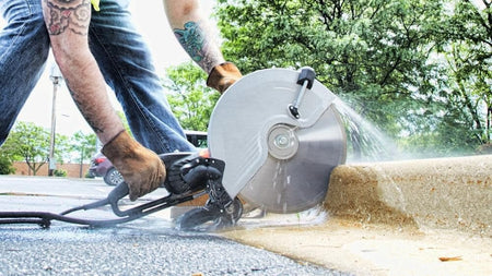 Worker using a large circular saw to cut concrete curb outdoors; water spray visible, tattoos on arms, with trees and building in background.