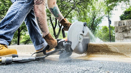 Worker cutting a concrete curb with a power saw and water spray; wearing gloves and boots, tattoos visible, with trees and stone wall in background.