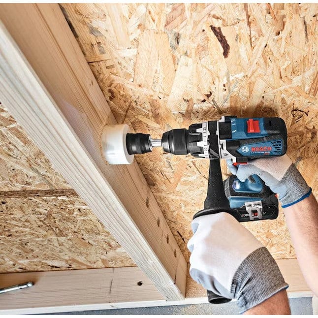 Worker using Bosch cordless drill with hole saw to cut a circular hole in OSB wall, wearing gloves and working on a framed structure.
