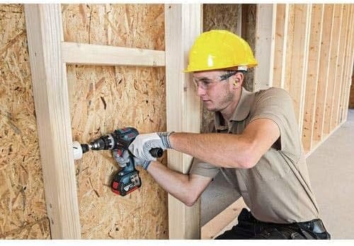 Person wearing safety gear using a power drill with hole saw attachment to bore into a wooden wall frame on a construction site.