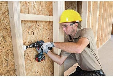 Person wearing safety gear using a power drill with hole saw attachment to bore into a wooden wall frame on a construction site.