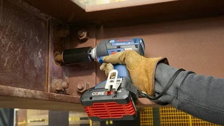 Person using a Bosch CORE18V cordless impact wrench to tighten a bolt on a steel beam in an industrial setting, wearing protective gloves.