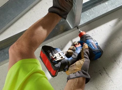 Worker using a Bosch CORE18V cordless drill to fasten a screw into a metal frame, wearing gloves and a bright yellow-green shirt.