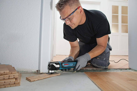 Person using Bosch GOP40-30 multi-tool to trim wood flooring near a doorway, wearing safety gear during a home renovation project.