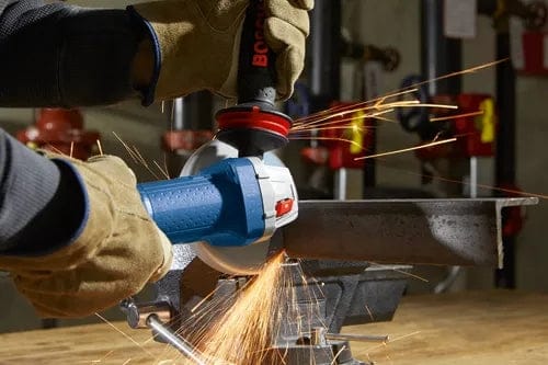 Person using a Bosch angle grinder to cut metal on a workbench, with visible sparks and protective gloves, demonstrating active use in a workshop setting.