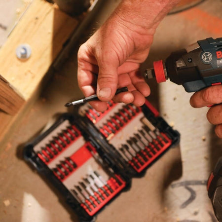 Person using a Bosch power drill near two open bit cases containing organized screwdriver and drill bits; construction setting with wood beams and concrete floor.