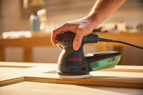 Close-up of hand operating Bosch electric sander on wood surface, actively sanding in a workshop with visible tools and workbench.