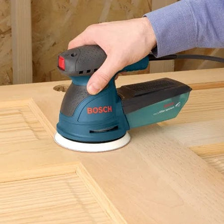 Person using Bosch 5-inch random orbit sander on a wooden door panel, smoothing the surface in a workshop setting with wood paneling in the background.