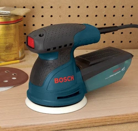 Bosch random orbit sander placed on wooden surface next to sanding disc and wood finish can, showing microfilter dust system and workshop pegboard background