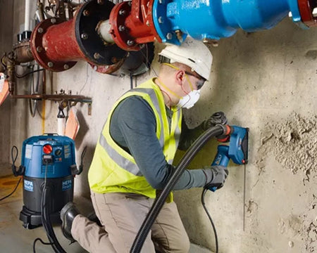 Construction worker using a power tool with HEPA vacuum system to drill into concrete wall; worker wears full safety gear including hard hat, mask, and high-visibility vest in industrial setting.
