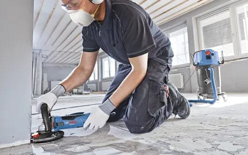 Person kneeling and grinding a concrete floor during renovation, using a handheld tool with vacuum hose attachment in a well-lit modern interior.