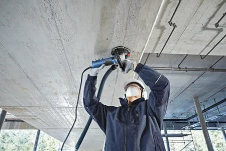 Worker using a power tool with dust extraction to grind the underside of a concrete ceiling, wearing protective gear in an industrial construction setting.