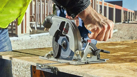 Worker in high-visibility vest using a circular saw to cut plywood at a construction site, with blade visibly engaged in active cutting.