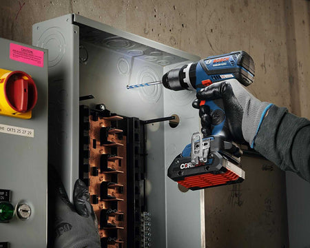 Worker using Bosch cordless drill on metal frame of an electrical panel; wearing gloves with copper bus bars and control buttons visible in the background.